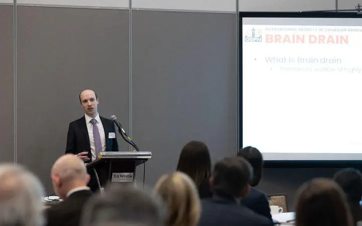 Thomas is speaking at a in front of a projector screen saying 'Brain Drain' at Research Canada's annual stakeholder action roundtable.